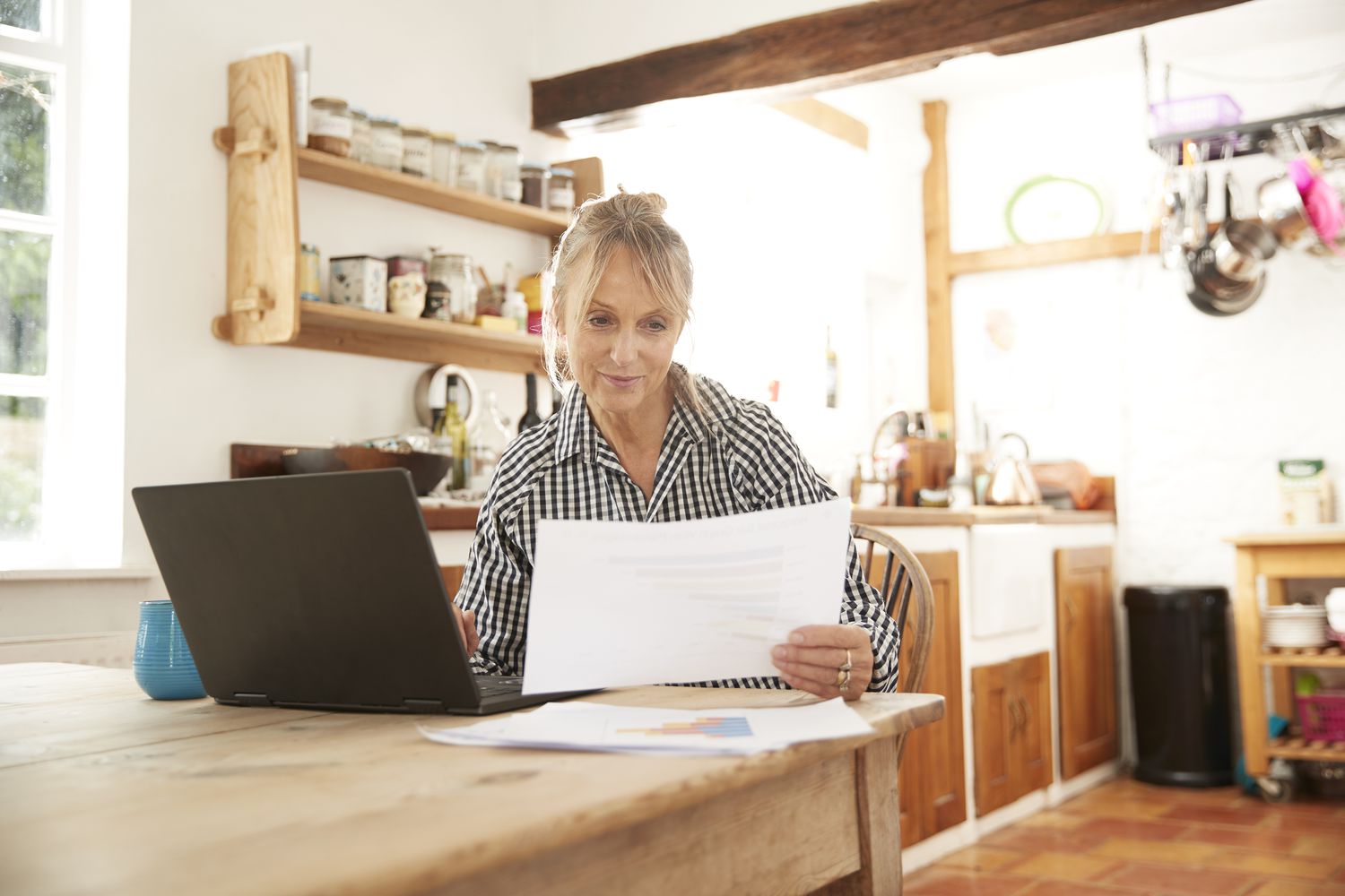 Woman in her 50s sitting at her kitchen table and looking at financial documents and a laptop