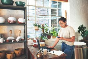 A person using a phone while standing by a desk in a room with pottery items and potted plants