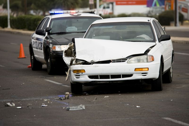Police car parked behind white sedan that's been in an accident, with serious passenger-side damage and broken glass and plastic around it.