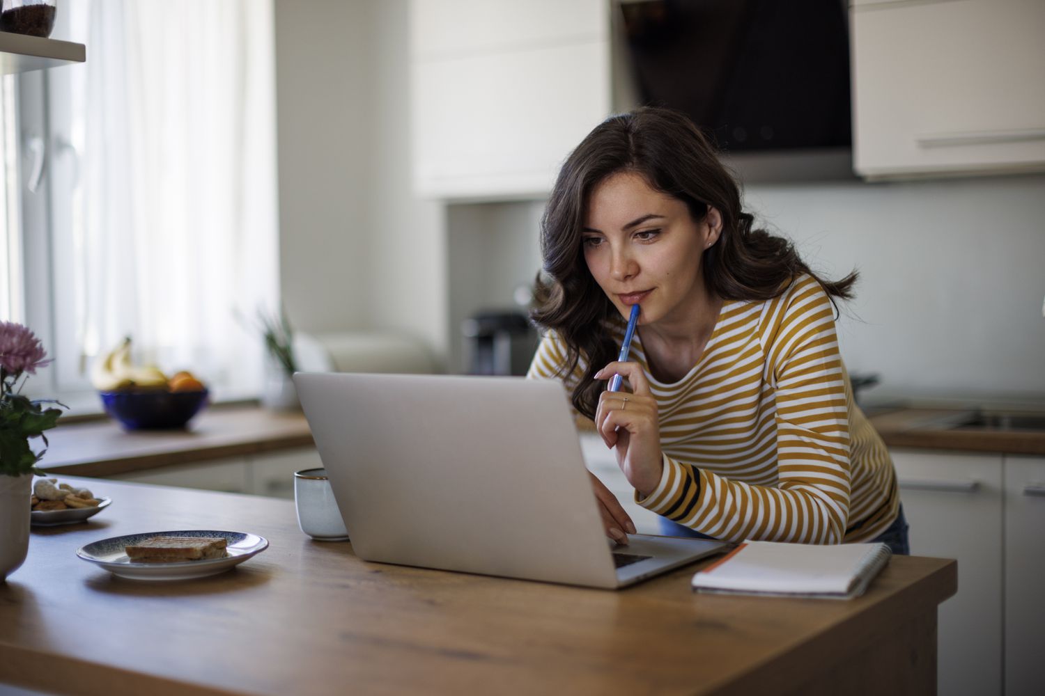 A woman with brown hair and a striped shirt stands at a kitchen counter with a laptop, pen, and paper