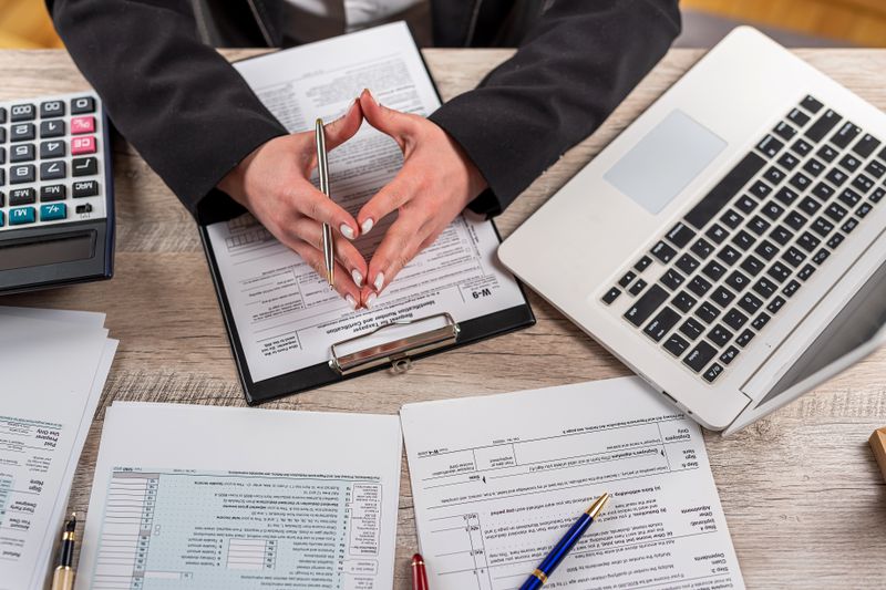 A tax accountant has various tax documents, a calculator, and a laptop spread on their desktop as they calculate taxes for a wealthy client.