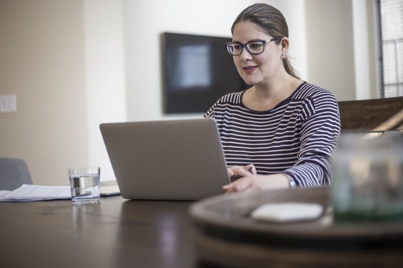 A woman with glasses in a striped shirt sits at a table with a laptop and a glass of water