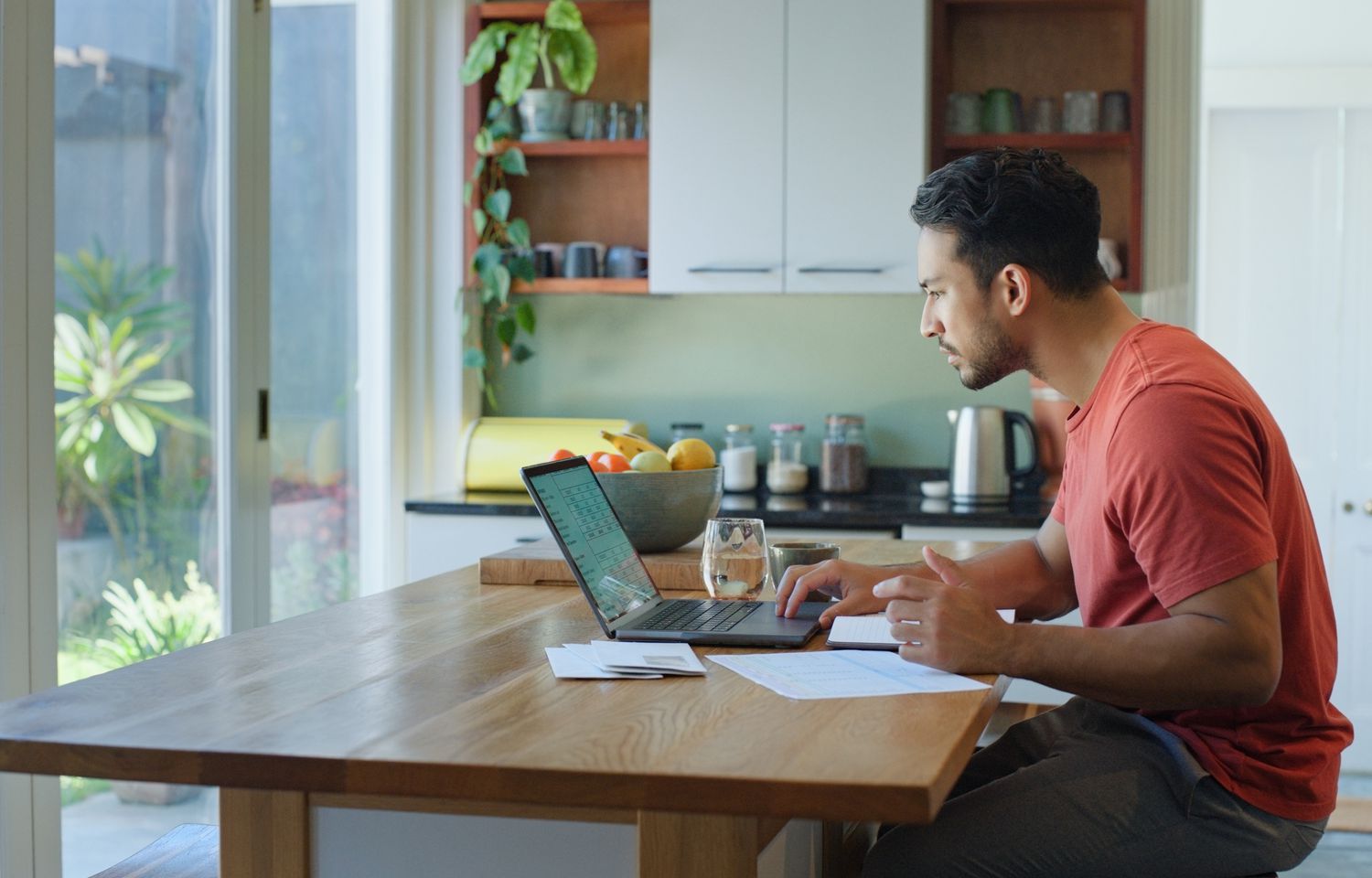 A man in a red t-shirt sits at a kitchen table with a laptop and papers