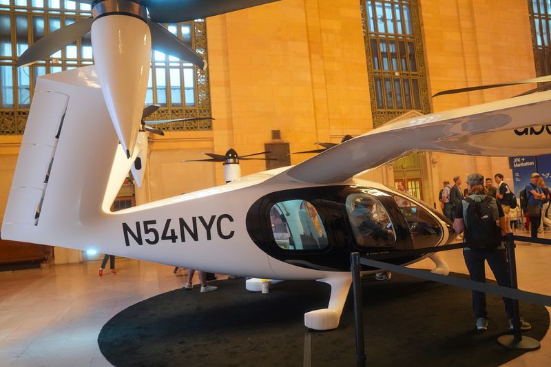 People stand around looking at a white and black Joby electric air taxi helicopter inside a building.