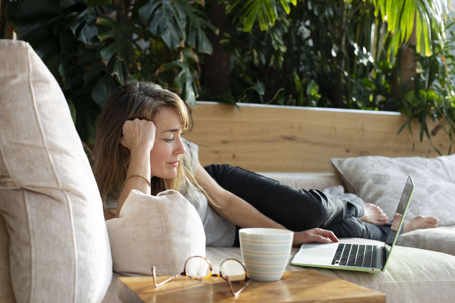 A woman lays on a couch with a laptop, mug, and glasses
