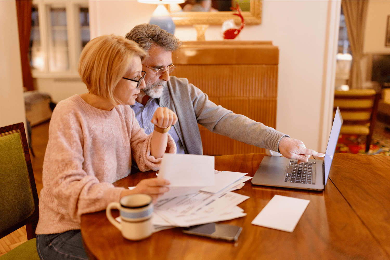 Older couple looking together at laptop and financial papers while sitting at dining room table
