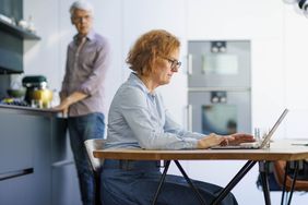 Woman working on a laptop while a man is looking at her from the kitchen