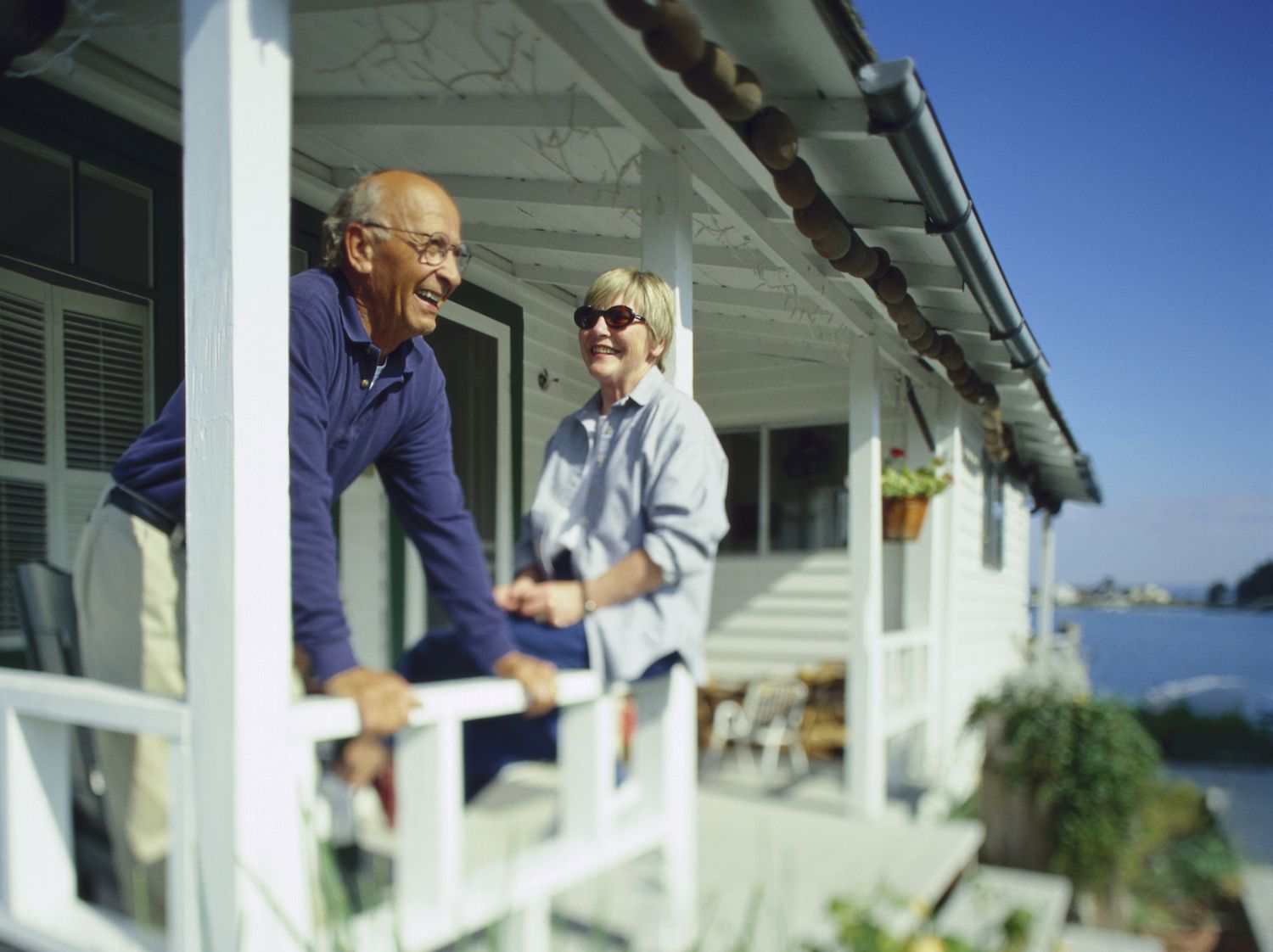A man and a woman stand and sit on a porch of a beach house looking out to the water