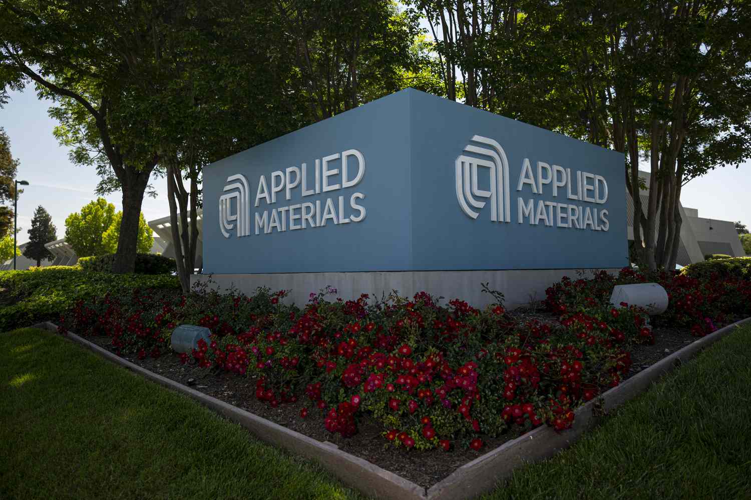 The blue Applied Materials sign is seen above a flowerbed with trees in the background.