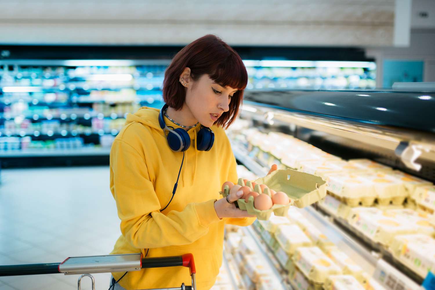 Young woman doing groceries in a local supermarket, holding an egg carton and checking the quality of the eggs before she buys them