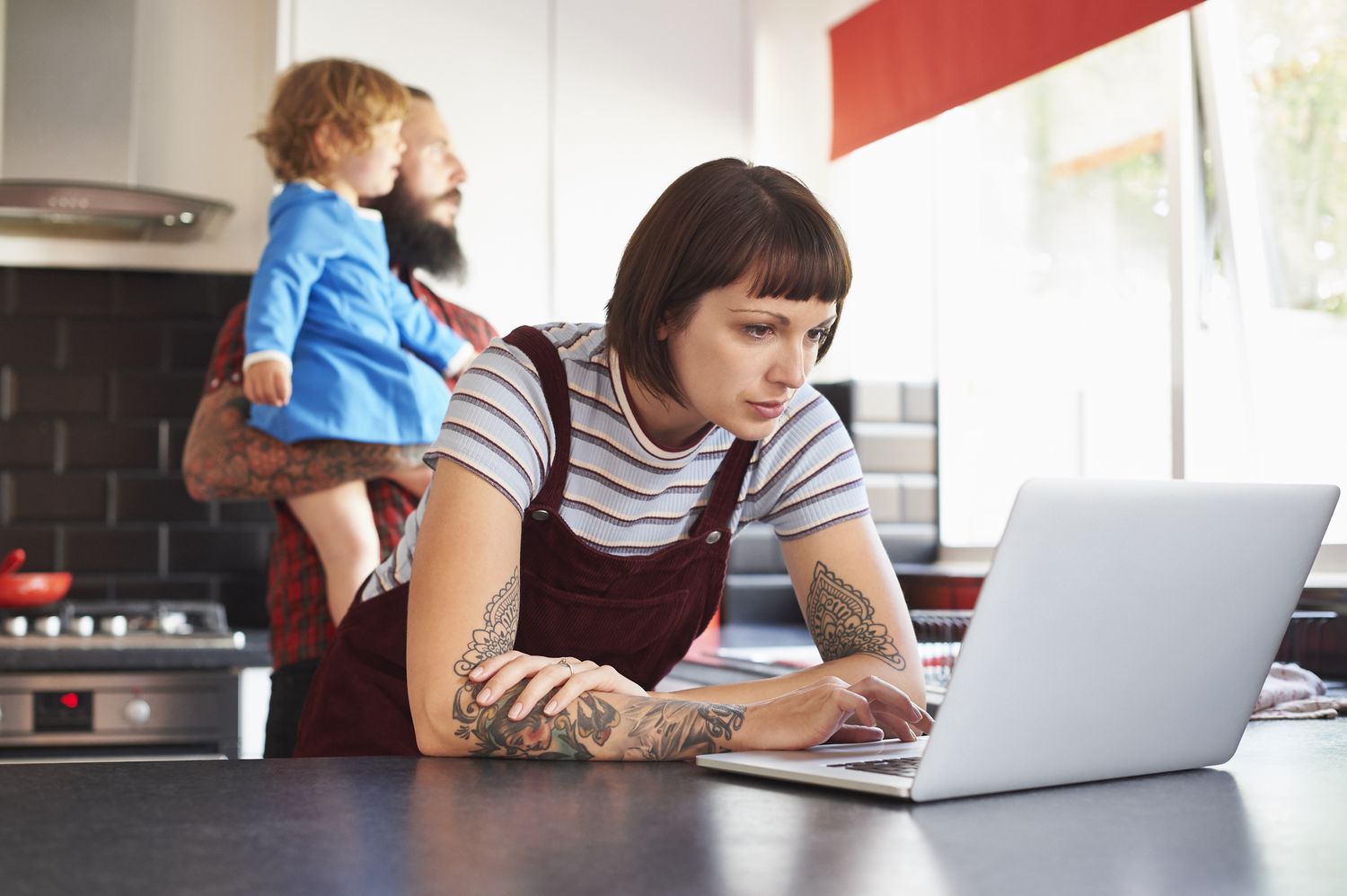 Young woman in her kitchen, looking at bank rates on her laptop while her partner and child stand in the background