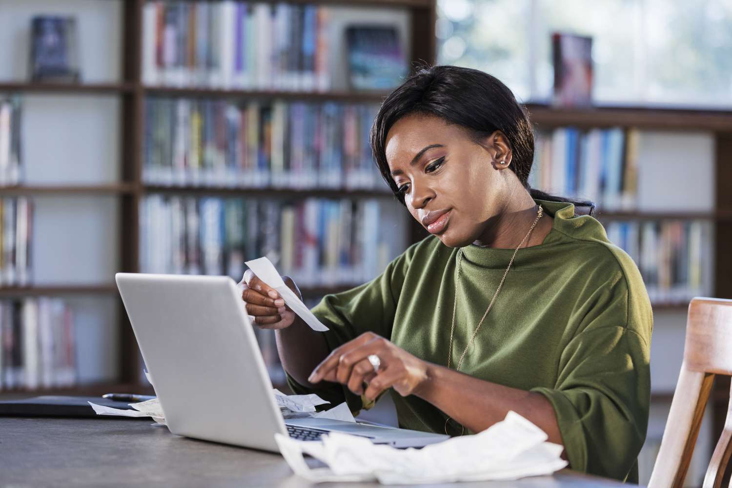A young woman sits at a laptop with a pile of receipts. She is holding one and about to start typing on the keyboard. 