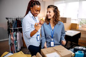 Two women work in a small business with boxes and clothing racks as they smile at each other and prepare a gift box.