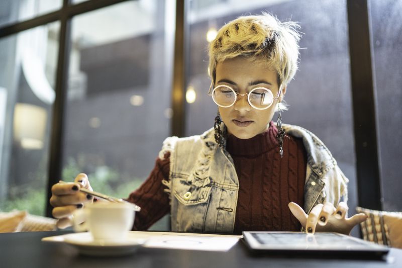 Young woman checking investments on a tablet while sitting at a coffee shop table. She has short blonde hair, round glasses, and long earrings, and is dressed stylishly.