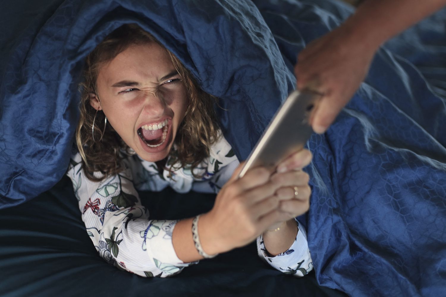 A hand reaches into the frame to take an iPhone away from an upset teenager under their covers in bed.