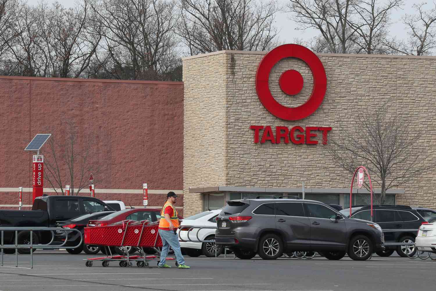 A Target worker pulls a group of shopping carts through a parking lot, with several cars and a Target store visible in the background