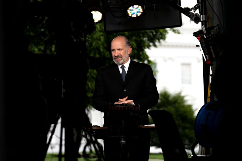 Howard Lutnick stands with his finders interlaced in front of his stomach. Cameras and lights surround him on the White House lawn.