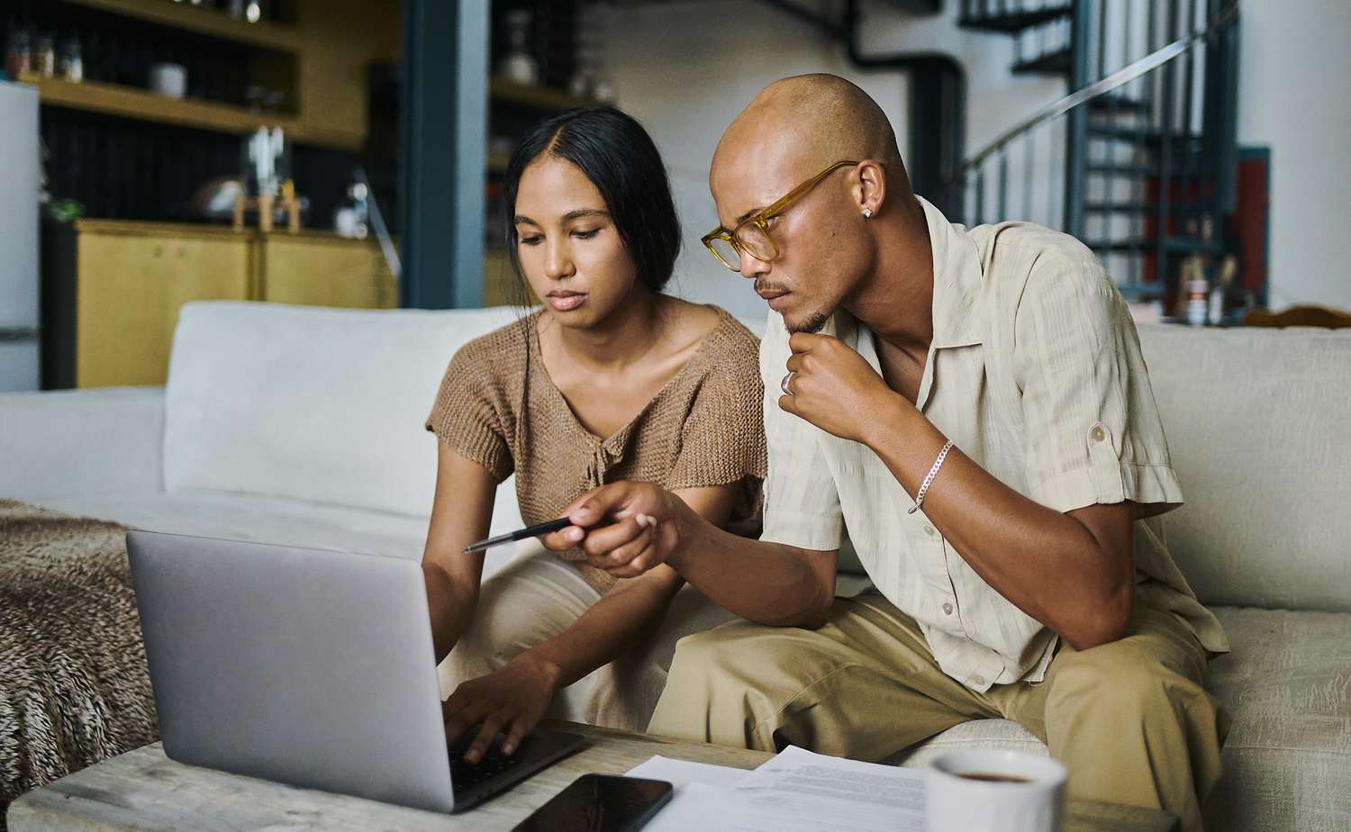 Two people sitting on a sofa, looking at a laptop together and holding a pen, appearing to discuss something