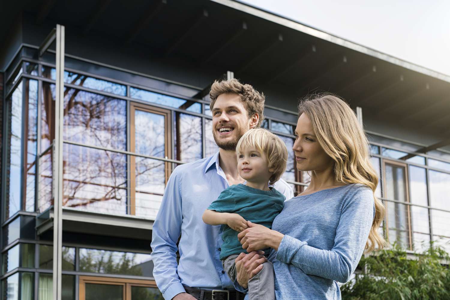 Smiling family of father, mother and toddler boy standing in garden in front of modern design house with glass facade.