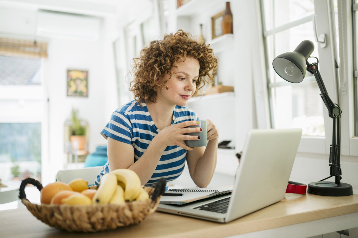 A woman stands at a desk looking at a laptop holding a mug