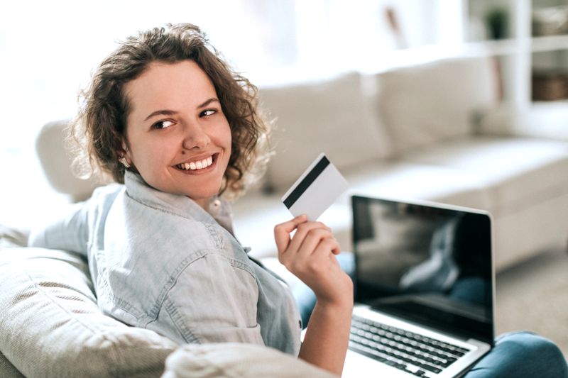 A woman sits on a couch with a laptop, happily holding up a bank card.