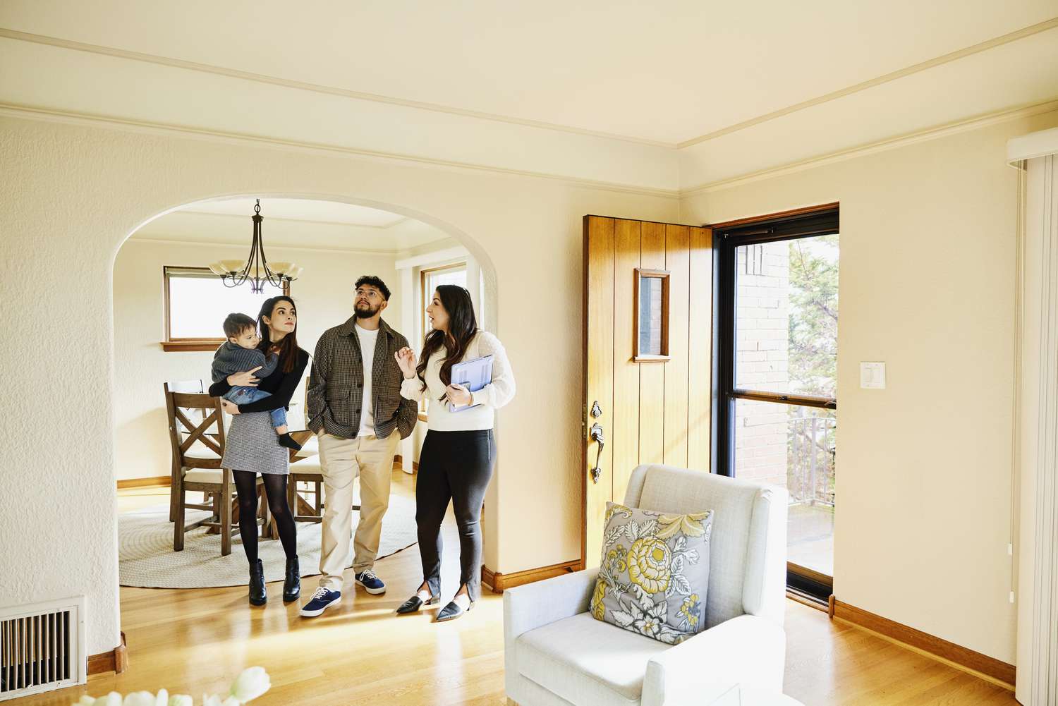 A woman holding a toddler and a man walk through a dining and living room of a house with a female agent.