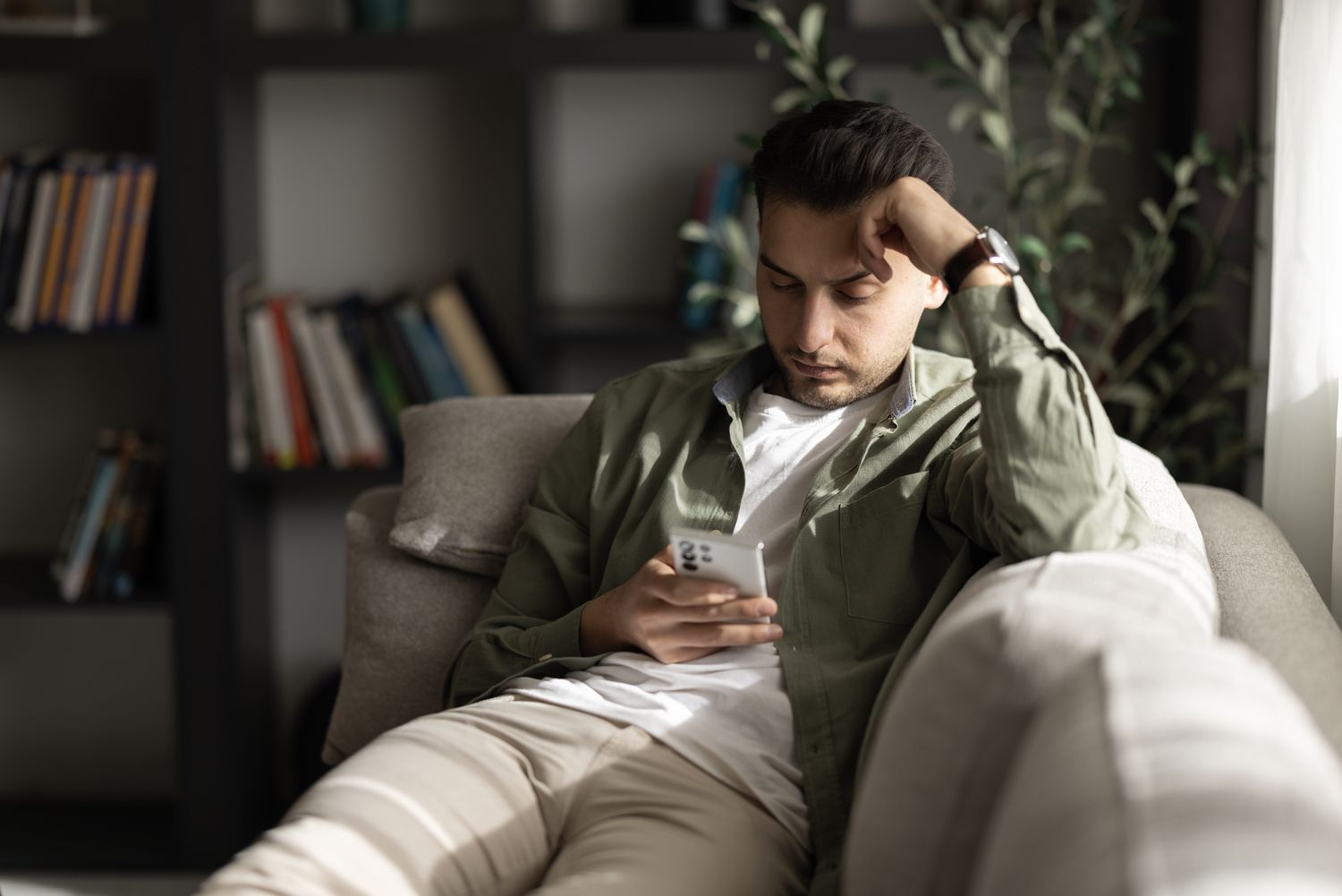 A man in a green shirt and tan pants sits on a couch looking at his smartphone