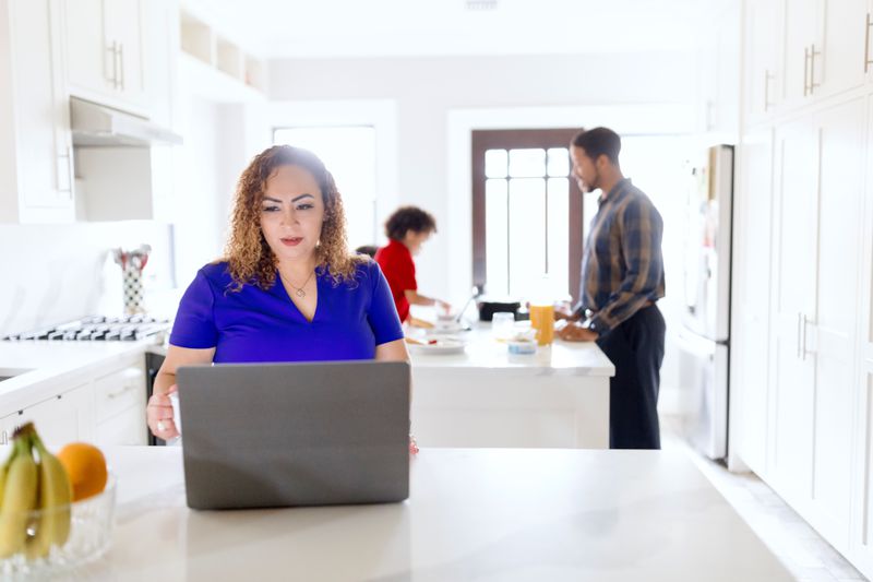 Woman in her kitchen looking at a laptop, while dad and son enjoy breakfast in the background