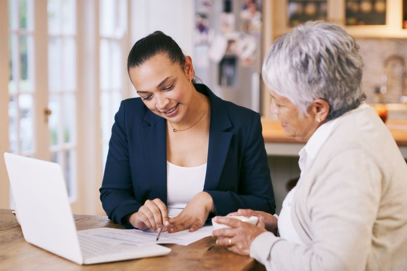 An older woman sitting at a kitchen table with a younger woman. There is a laptop and paperwork on the table that they are both looking at. 