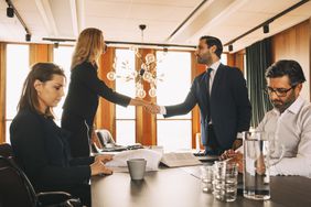 Two people shake hands over a conference room table where a man and woman sit not looking at each other. 