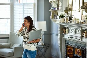 Woman in a living room listening on the phone while holding an open laptop.