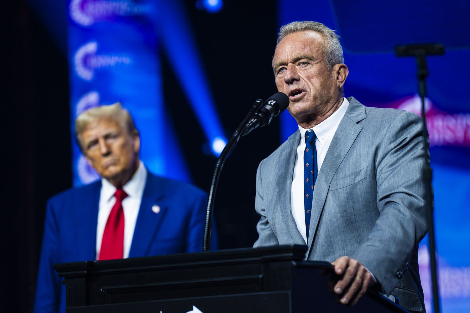 Robert F. Kennedy Jr. speaks at a podium while Donald Trump stands behind him in front of a blue background.