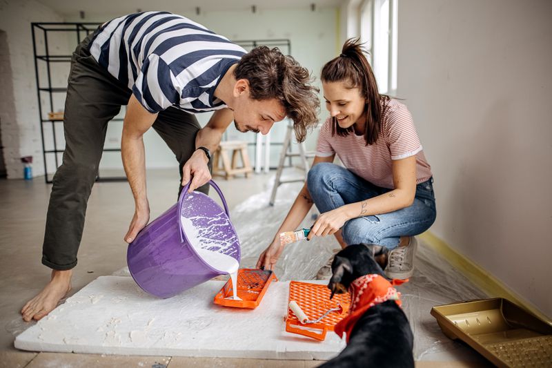Young couple mixes the paint to fix up the wall of their house with their dog standing and observing.