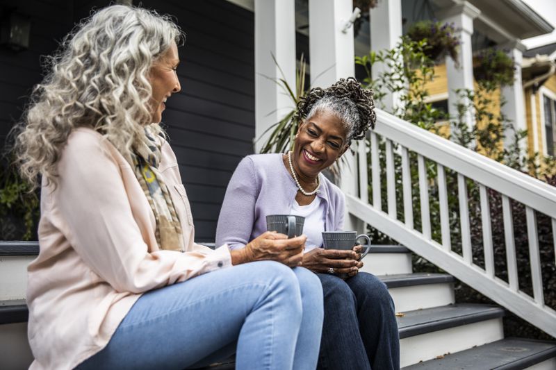 Two older woman sitting on the porch of a suburban home laughing and drinking coffee.