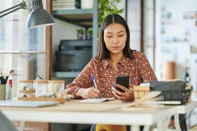 Young woman sitting at her home desk and looking seriously at her phone, with pen and paper in hand