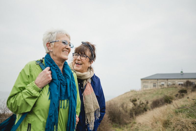Two older women walking on the beach together during the winter time, they are wearing warm clothing.