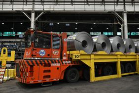 An orange truck is seen with rolls of steel on a trailer behind it.
