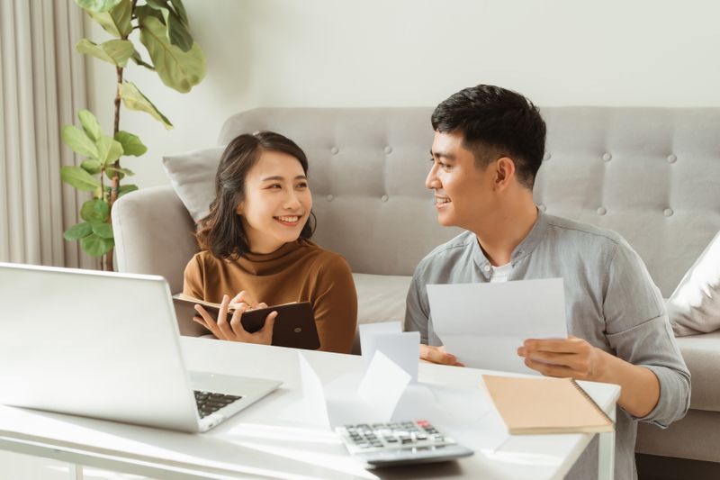 Two people discussing their finances using a notebook, laptop, and some papers while sitting in front of a couch.