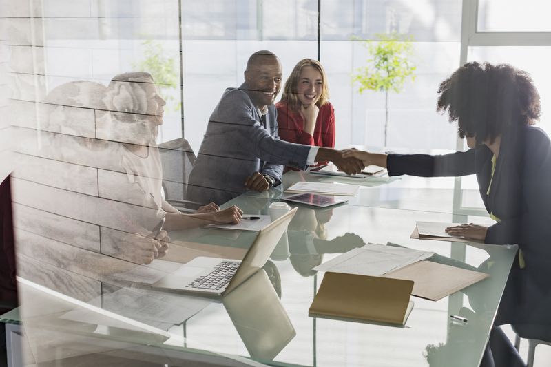 A businessman and businesswoman handshaking across conference table in meeting.
