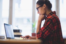 A person sits at a desk, working on a laptop. A pile of books is seen in the background.