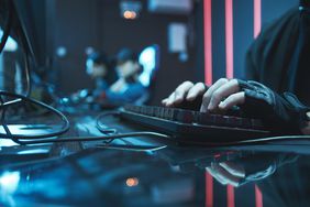 Close-up of someone's hands typing on a keyboard in an e-sports or gaming setup, blurred background shows players