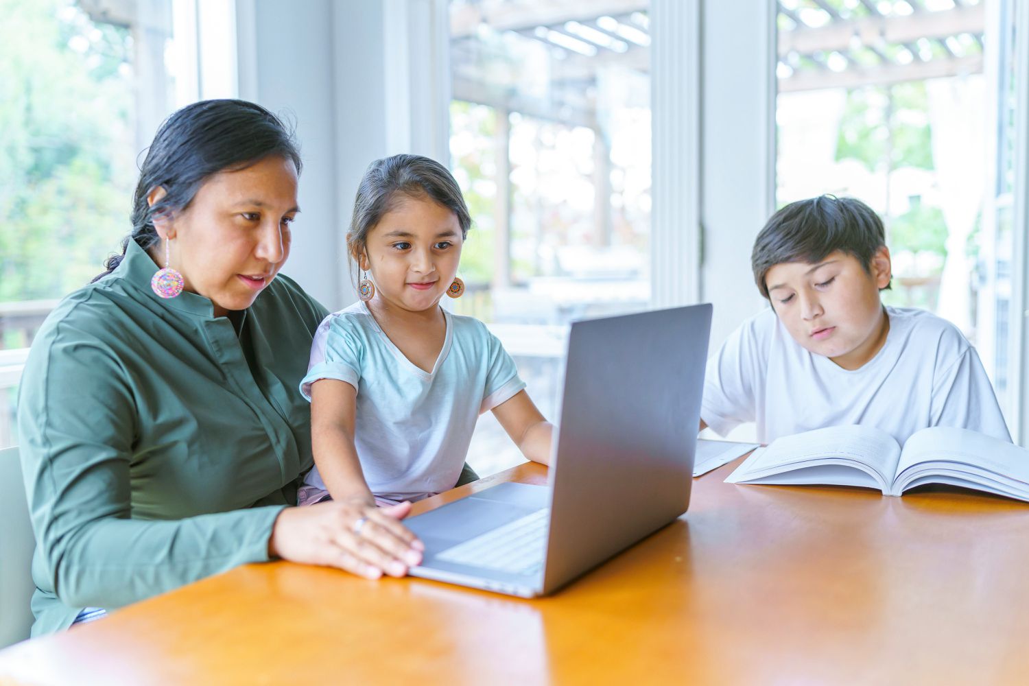 A Native American woman sits at a dining room table with her son and daughter while she works on a laptop computer.