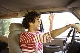 A happy, smiling man drives in a car with his hand out the window.