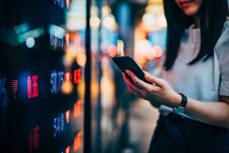 Businesswoman checking financial trading data on smartphone by the stock exchange market display screen board