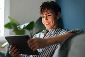 Young woman sitting on her sofa and looking happily at bank rates on a tablet