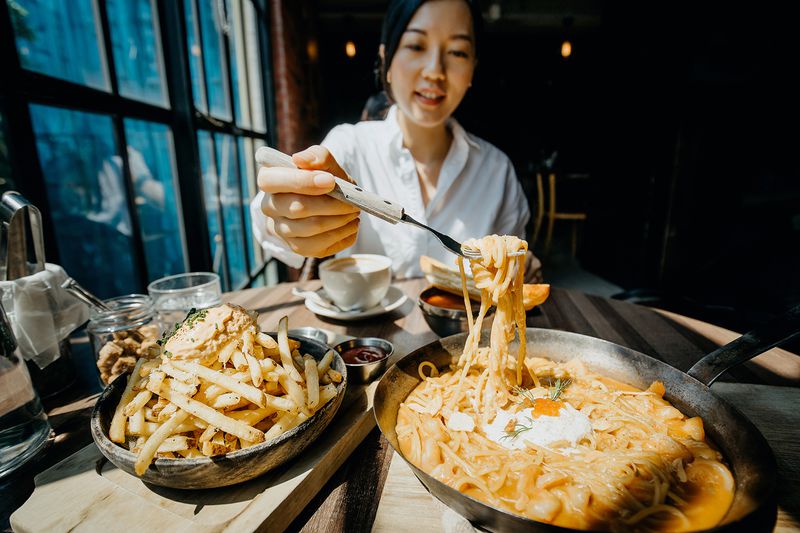 Young Asian Woman Sitting at a Table by the Window Enjoying the Warmth of Sunlight and Having Meal Joyfully in a Restaurant