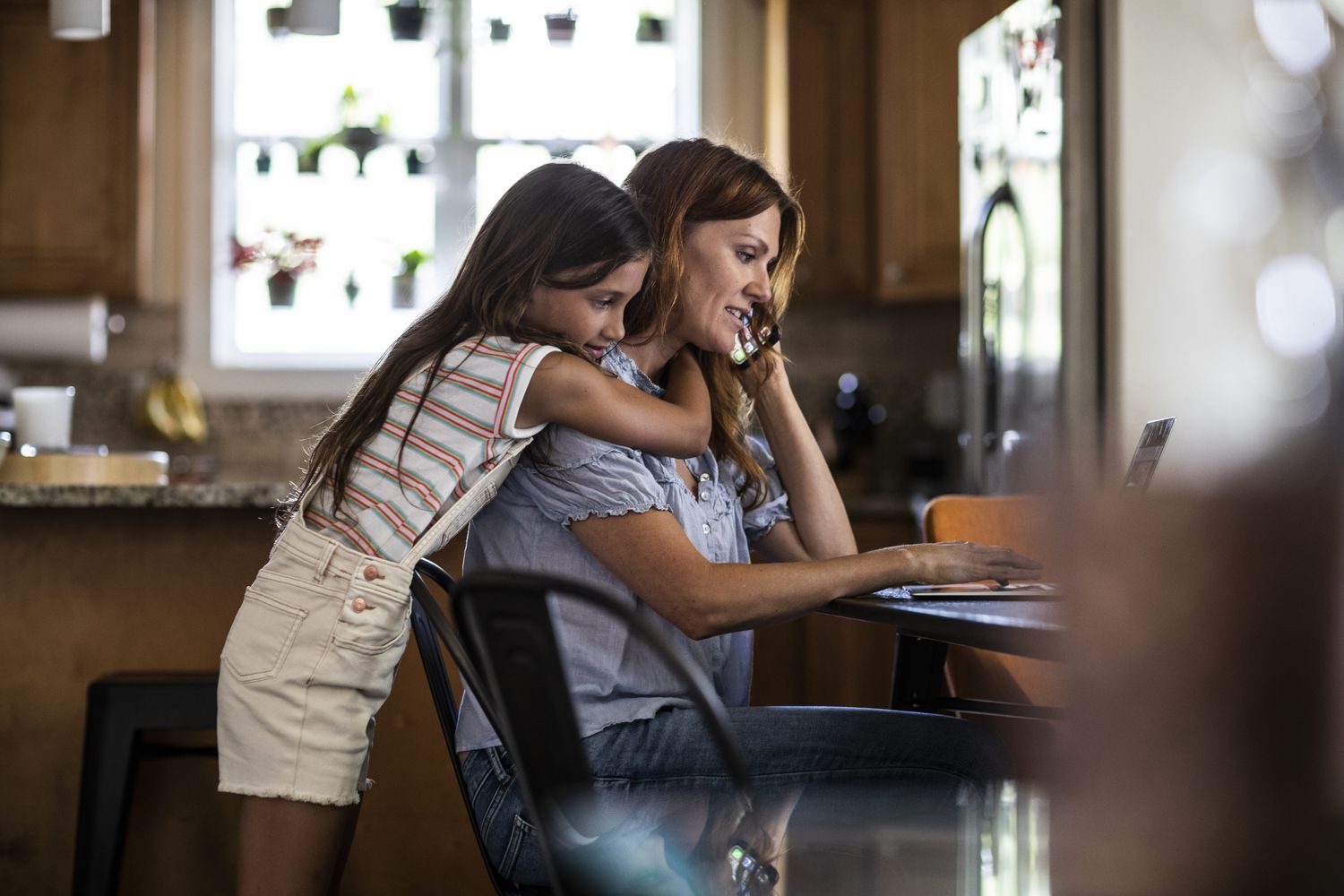 Daughter hugging mom from behind while the other one is on the phone looking for ways to save their house from foreclosure.