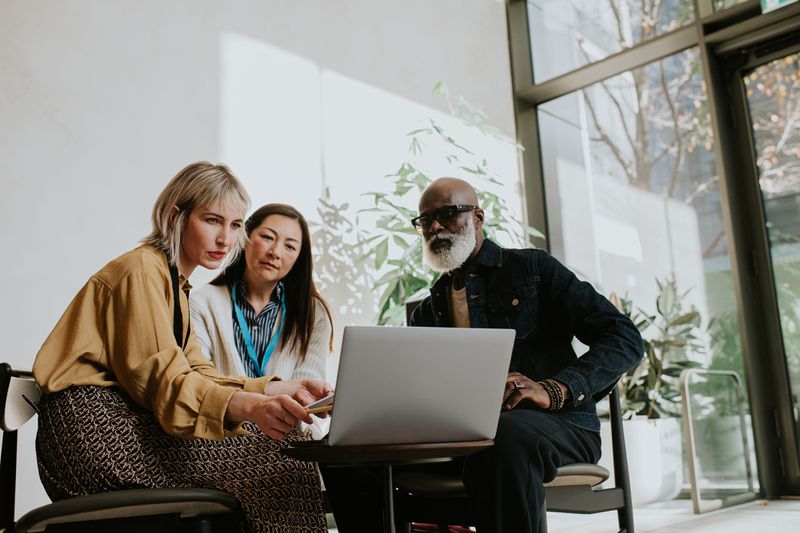 Three people sit around a laptop. One woman is presenting to the group