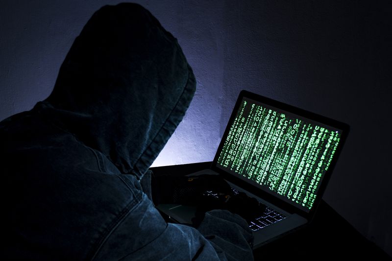 hacker in a hood whose face is concealed looking at a laptop whose screen is green binary numbers