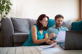 Couple looking at paper and laptop while sitting on the floor in front of their couch